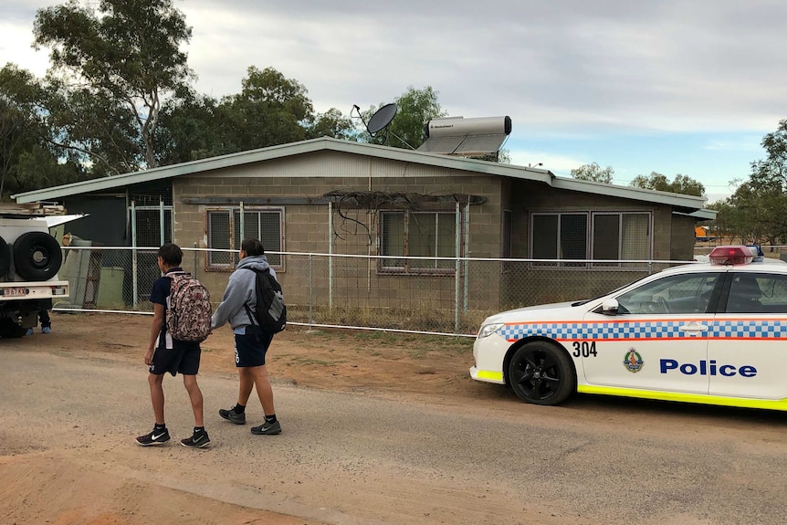 Two kids with backpacks walk past a house with a police car parked out front.