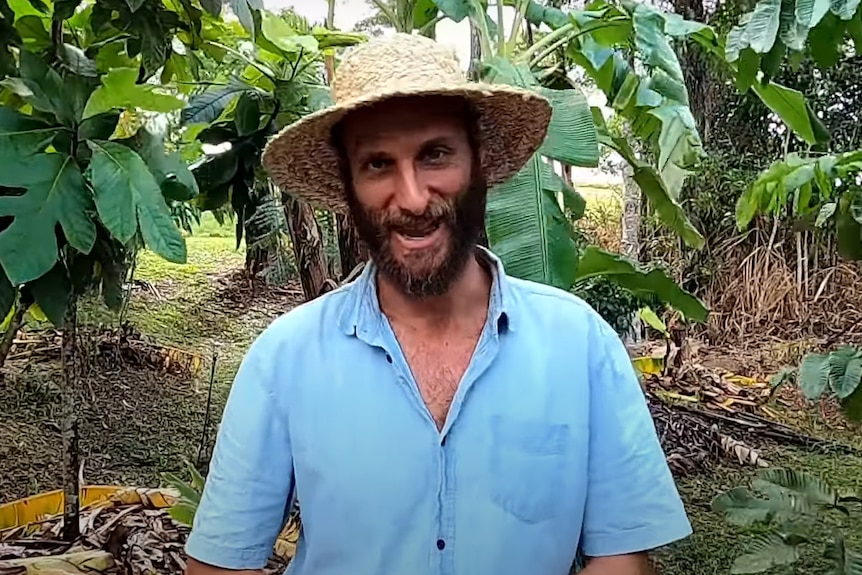 A bearded man wearing a broad-brimmed hat stands in a garden of fruit plants.