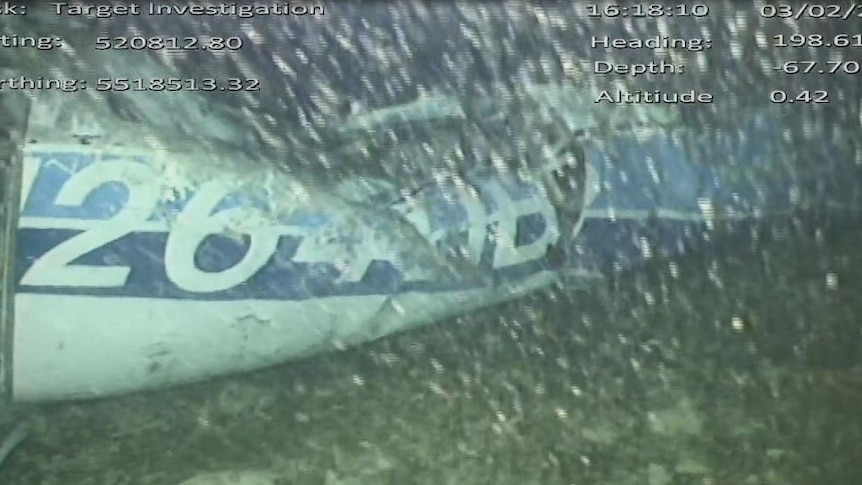 An underwater image shows a crumpled plane fuselage on the seabed lit with light from the camera as bubbles appear in foreground