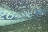 An underwater image shows a crumpled plane fuselage on the seabed lit with light from the camera as bubbles appear in foreground