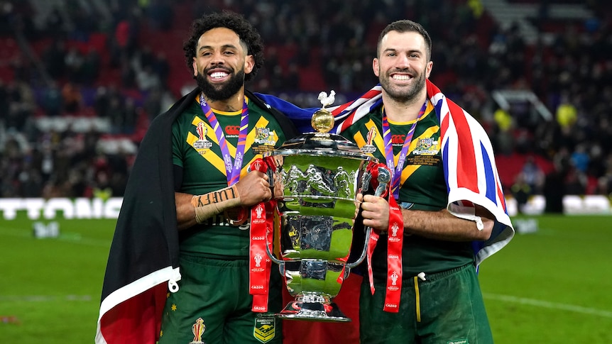 James Tedesco and Josh Addo-Carr hold the RUgby League World Cup trophy while draped in the Aboriginal and Australian flags.