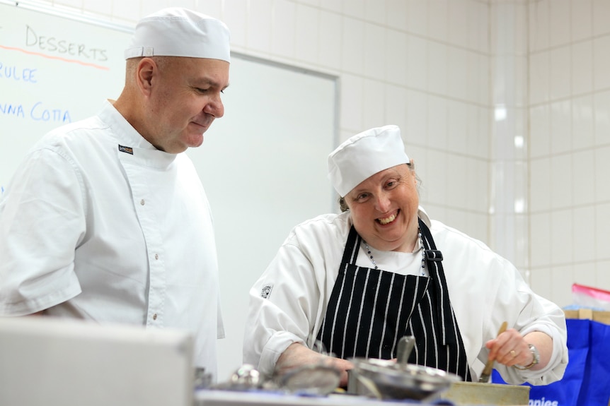 A lady in chef's clothes smiles as she works in the kitchen as she is watched by a man also in chef's clothes.