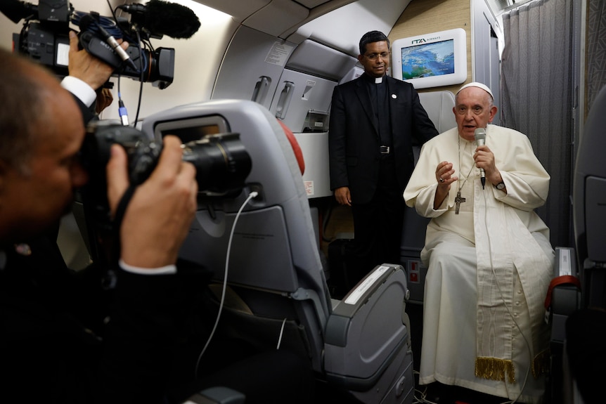 Papa Francisc stă în interiorul unui avion, cu un portret în prim plan și o persoană care stă în spatele lui. 
