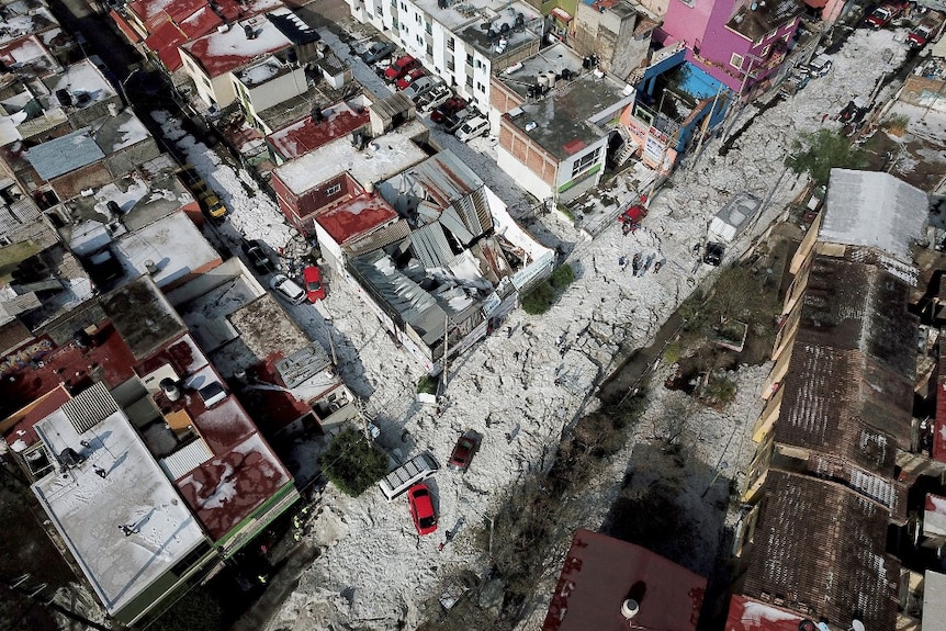 A birds-eye view of streets and houses. Some of the rooves are damaged, and the streets are covered in whiteness.