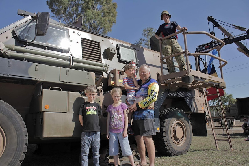 A man stands with his three children in front of a Bushmaster tank, his wife is on top of the vehicle wearing Army uniform.