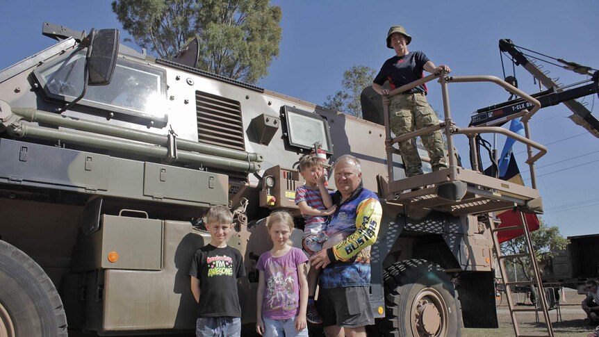 A man stands with his three children in front of a Bushmaster tank, his wife is on top of the vehicle wearing Army uniform.