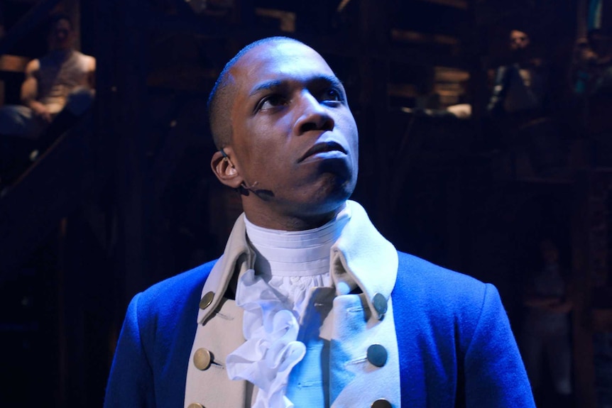Leslie Odom Jr, in a blue coat and frilly shirt, looks up and out