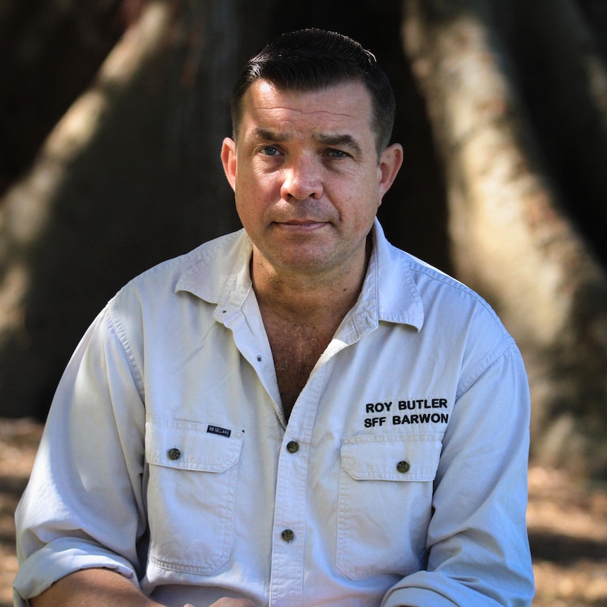 A man in a white-coloured shirt sits in the shade of a tree.