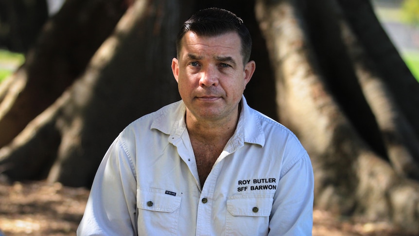 Caucasian man with dark hair sits in dappled light with white shirt.