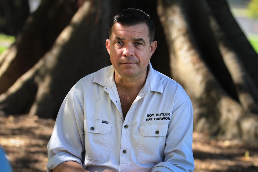 A man in a white-coloured shirt sits in the shade of a tree.