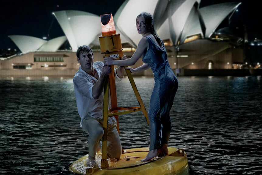 Sydney Sweeney in a blue dress and Glen Powell in a white shirt standing on a buoy in waters with Sydney Opera House in shot