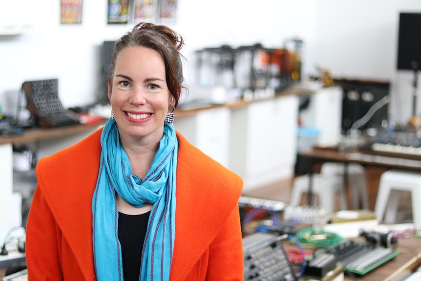 A woman in a bright organe jacket and pale blue scarf smiles.