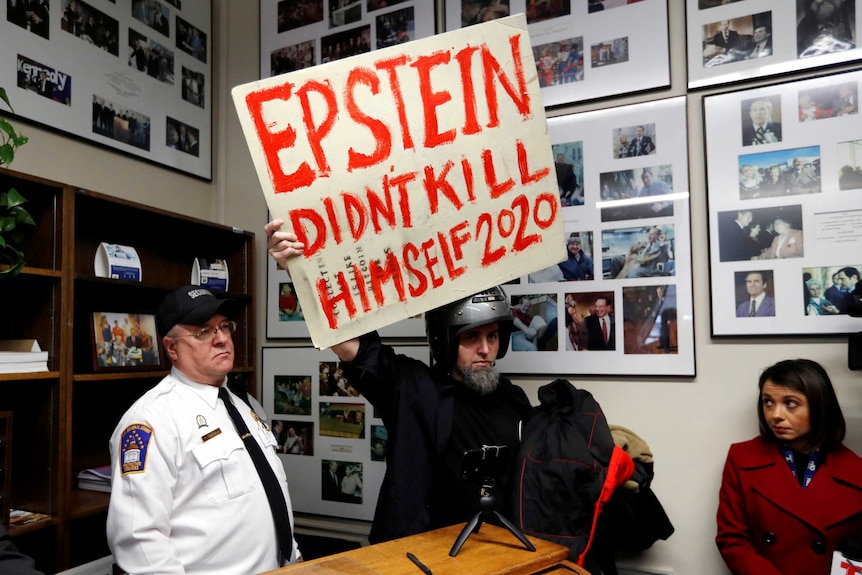 A man holding a sign and wearing a helmet in an office is curtailed by a security guard.