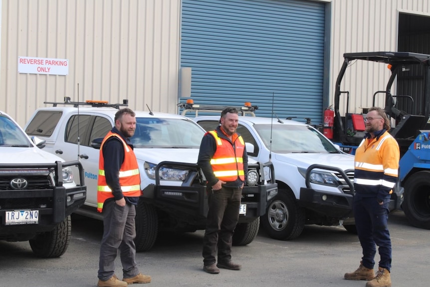 Three men in high vis stand in front of work cars. 