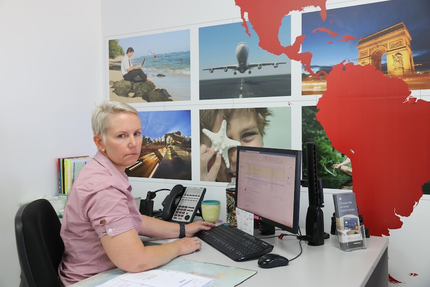 blonde woman in a red shirt sits at her travel agency office looking serious