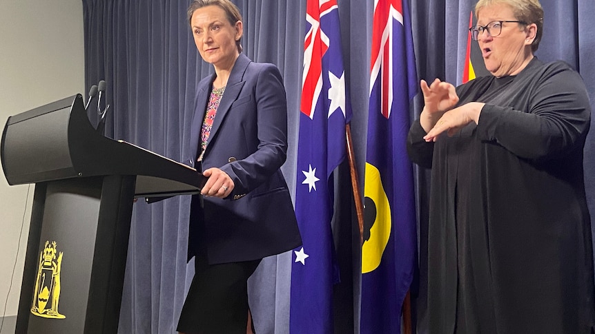 Amber Jade Sanderson speaking from a podium at COVID presser, with an Auslan interpreter by her side.