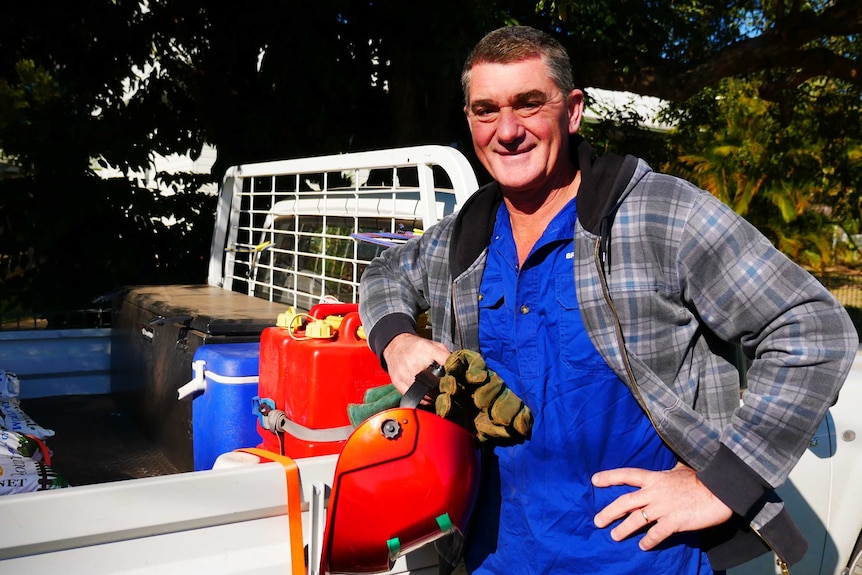 Middle aged tradie in perfect outfit: crumpled brightly coloured collard shirt, welding gloves and helmet, leaned up against ute