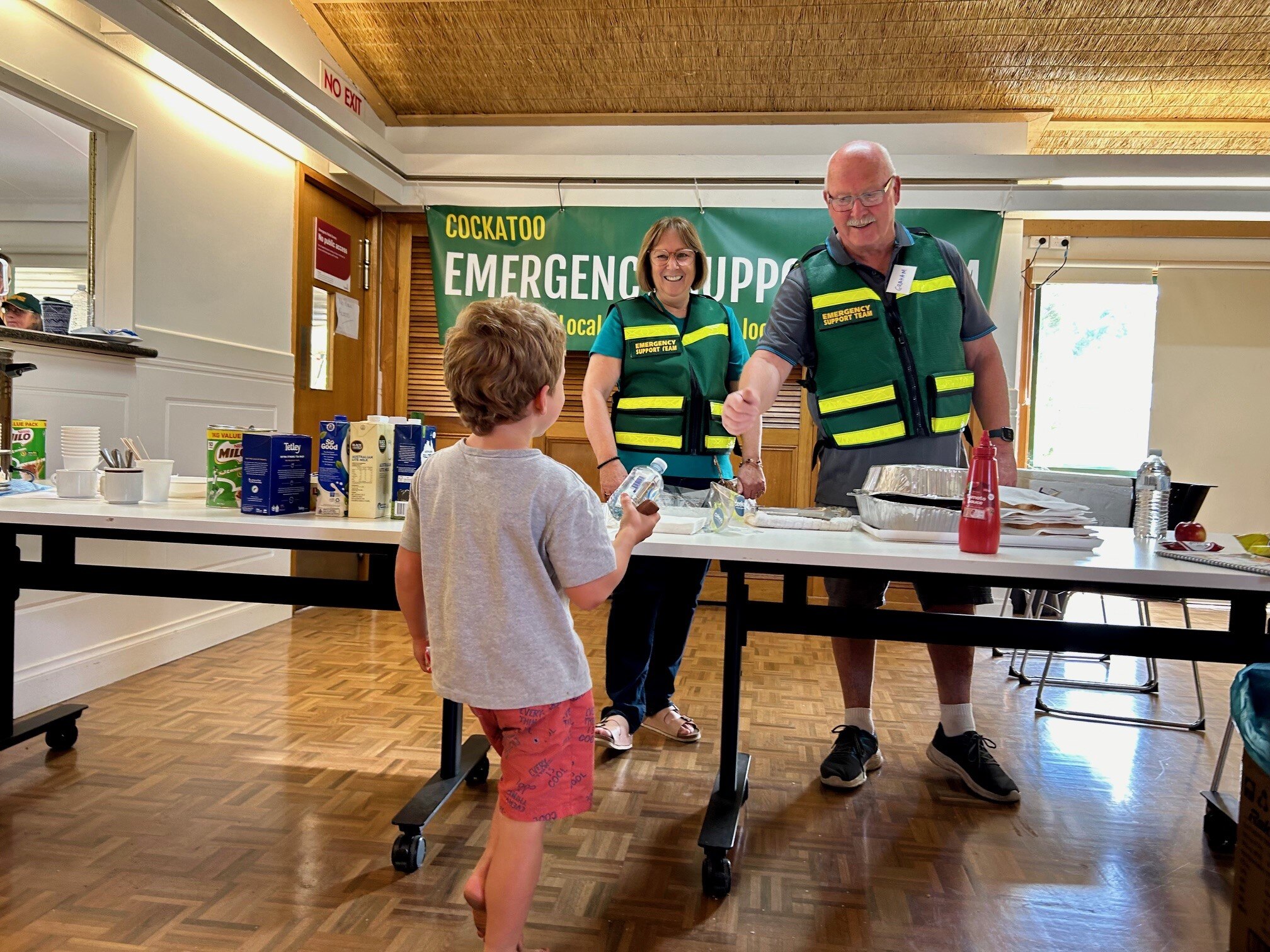 A young boy walks up to a table where a man and woman are serving food.