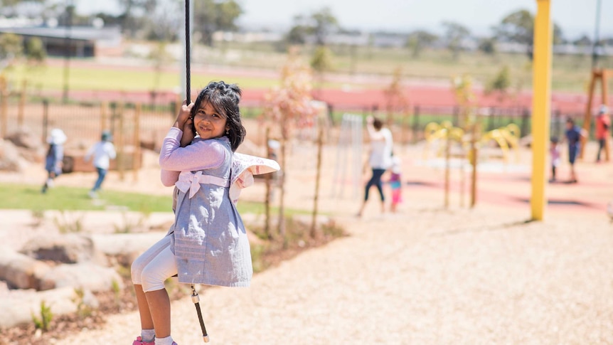 A young girl smiles at the camera while riding a flying fox, playground in background.