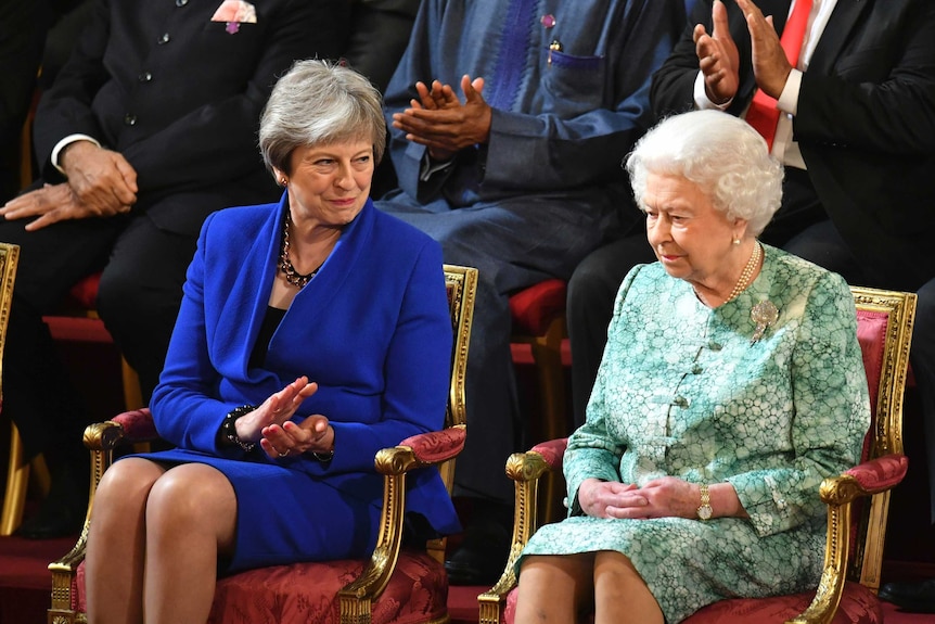 Queen Elizabeth II and Theresa May sit next to each other at the opening ceremony. May is clapping and looking at the queen.