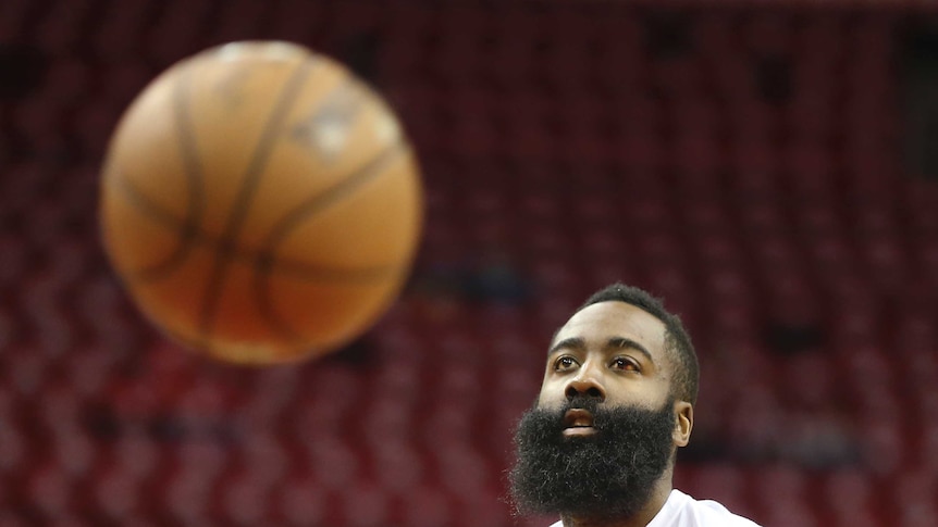 James Harden wearing a white shirt with a basketball in front of him.