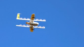 A delivery drone, with propellers spinning, hovers against a blue sky.