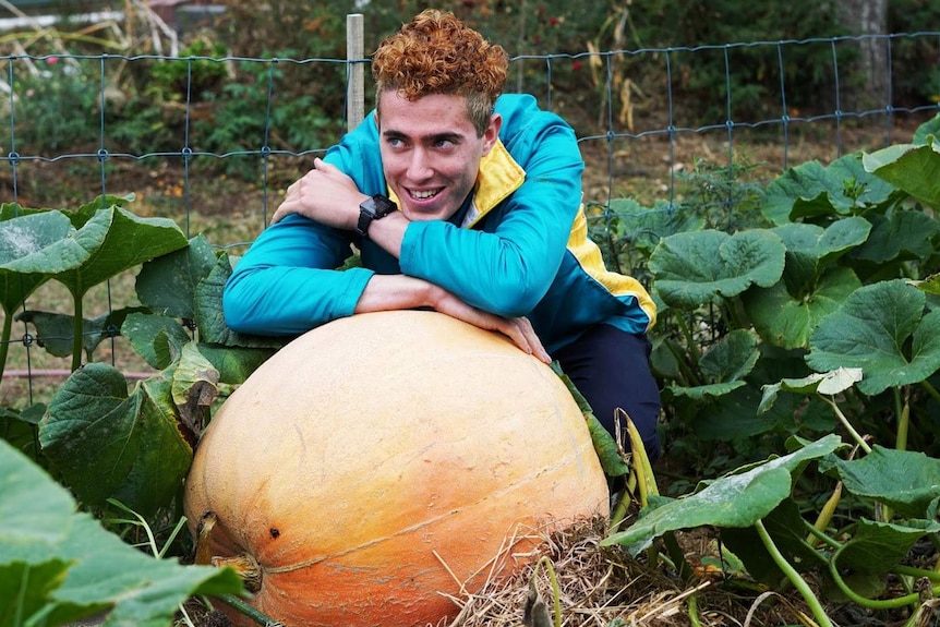 A young man with dyed orange hair stands with his arms folded over a giant pumpkin surrounded by large green leaves in a garden.