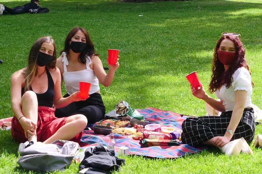 Three young women wearing face masks hold up cups happily in a park.