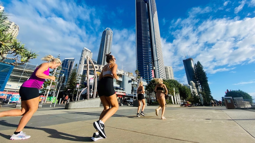Four women in active wear running along a concrete path with buildings in background.