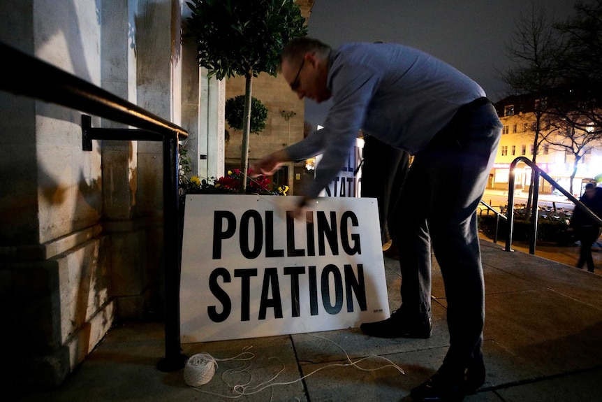 A man fastens a 'polling station' sign to a building in the dark