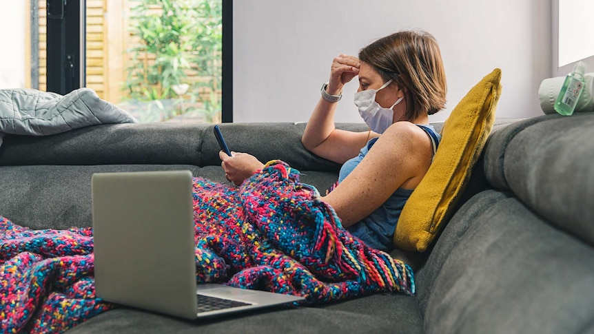 Woman self-isolating on the sofa wearing a mask and looking at her phone.