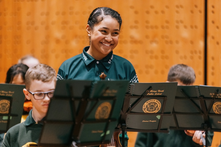 Cecilia Ikamanu, an 11-year-old Tongan Australian girl in a yellow and green school uniform smiles in front of a music stand