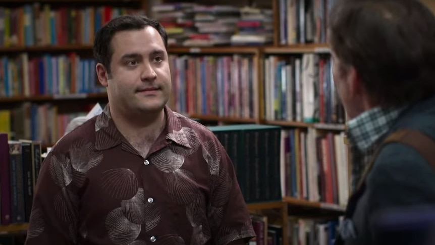 man with short dark hair and patterned shirt grinning in front of bookshelves and back of mans head
