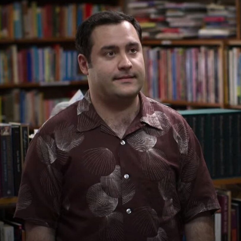 man with short dark hair and patterned shirt grinning in front of bookshelves and back of mans head