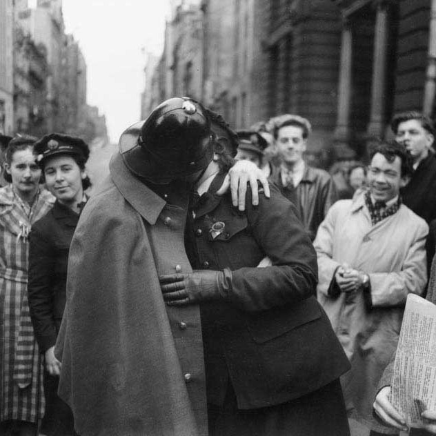 Black and white photo of a policeman and a woman in military uniform kissing in the street surrounded by grinning crowds.