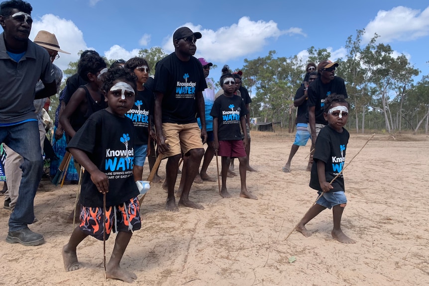 Two Aboriginal children with white ochre on their faces lead a group dancing in sand.