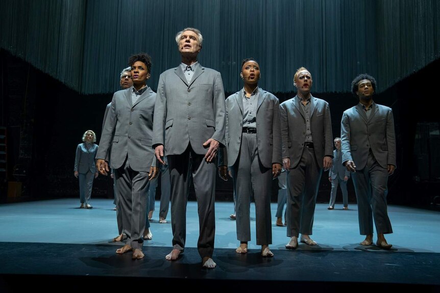 David Byrne stands on stage in front of a formation of 11 performers all barefoot and wearing grey suits, spot-lit, singing.