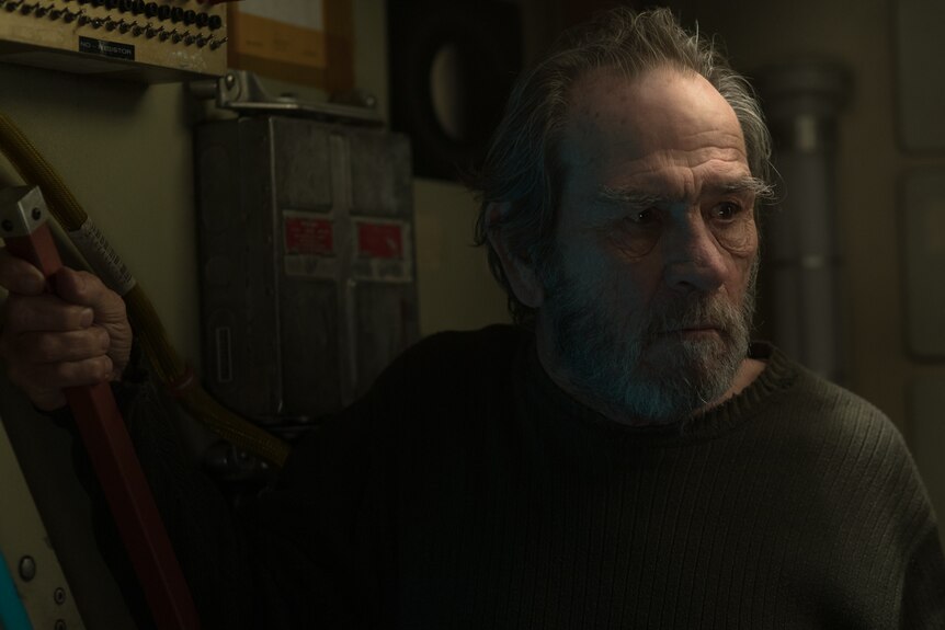 Darkened shot, interior, with older actor with beard and wearing dark green woollen jumper turned slightly sidewards.