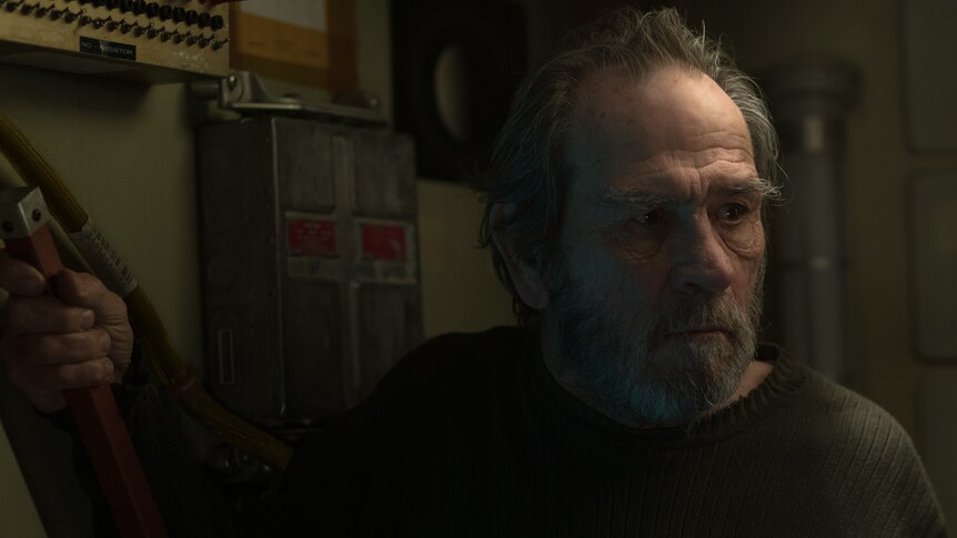 Darkened shot, interior, with older actor with beard and wearing dark green woollen jumper turned slightly sidewards.