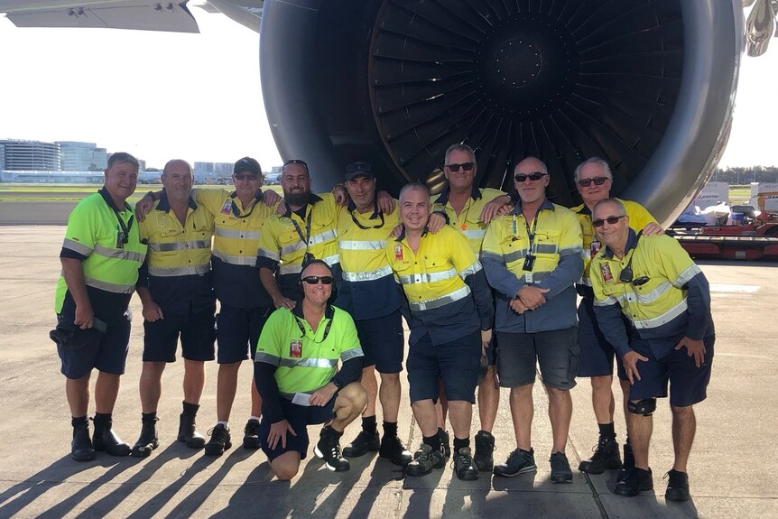 A group of 10 men wearing high vis shirts, navy shorts and boots stand in front of an aircraft's jet engine.