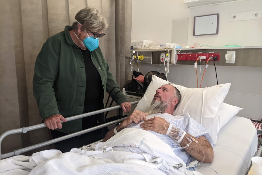 Grant Ottaway in a hospital bed looking up at a woman standing beside the bed.