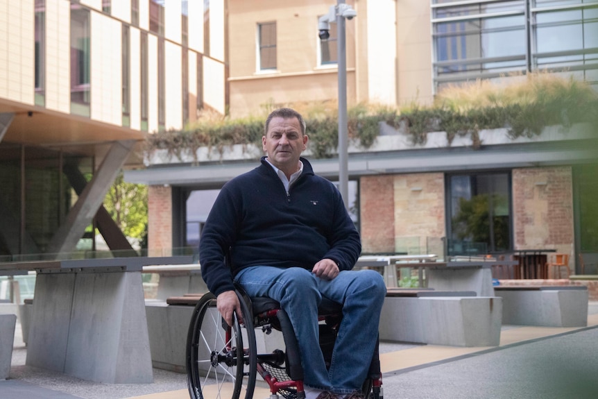A man sits in a wheelchair in front of bench seating with buildings behind.