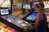 profile shot of a woman with short blonde hard and scarf headband, blue singlet plays pinball