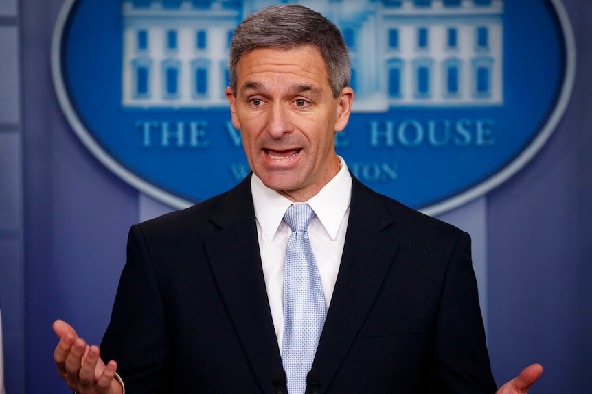 In front of the blue White House oval seal, Ken Cuccinelli speaks an gesticulates with his hands.