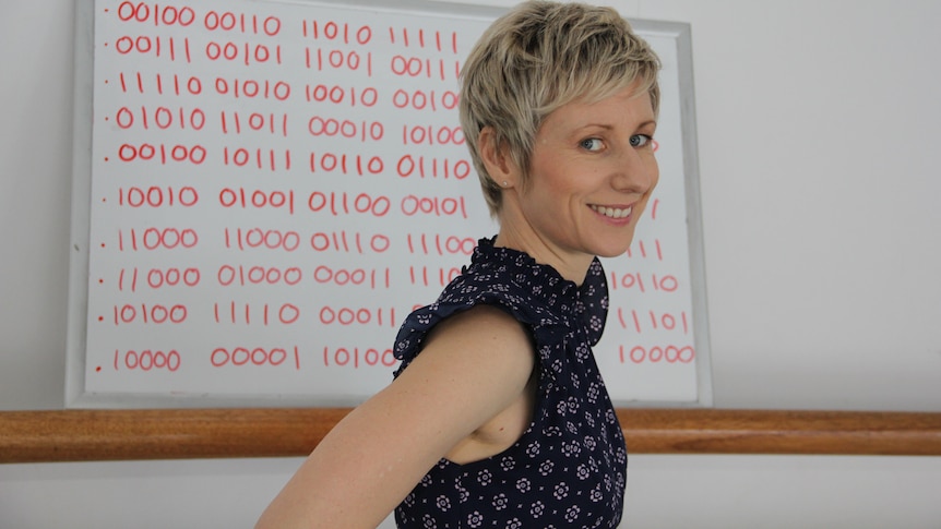 A smiling woman with short blonde hair stands in front of a whiteboard covered with hundreds of binary digits.