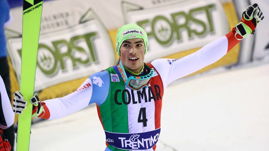 A smiling skier raises his fist in salute to the crowd while holding a ski in his other hand.