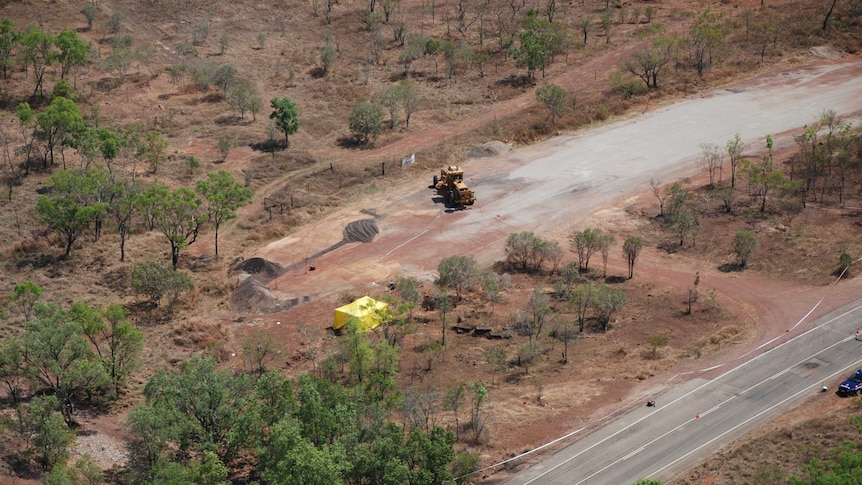 An aerial shot shows a road beside bushland. In a clearing, a grader is visible near a yellow tent.
