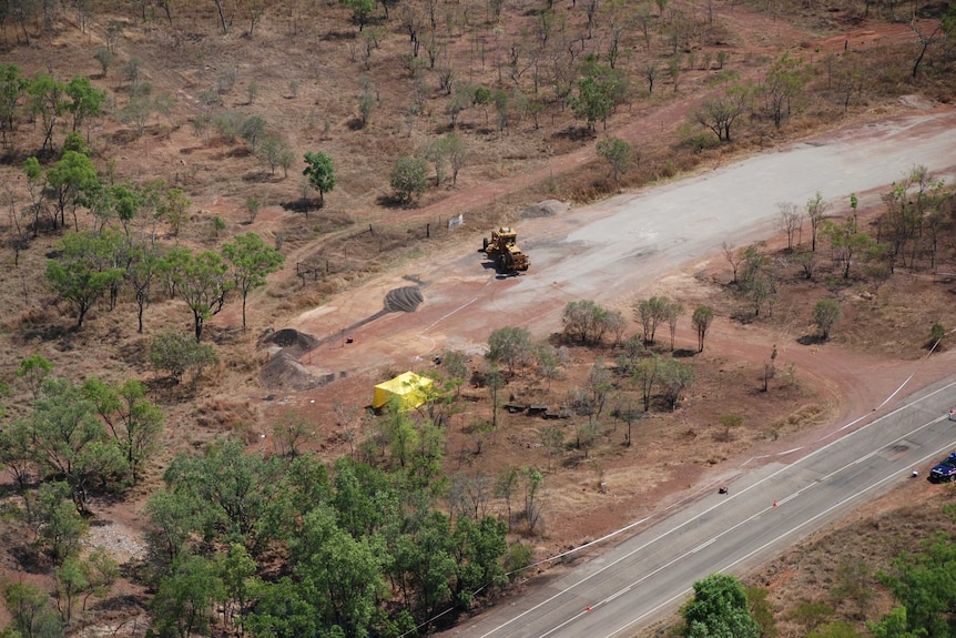 An aerial shot shows a road beside bushland. In a clearing, a grader is visible near a yellow tent.