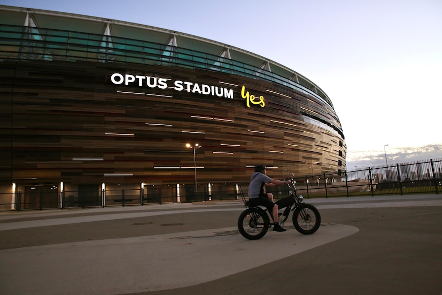 A man on a bicycle rides around the Perth Stadium as the sun goes down.