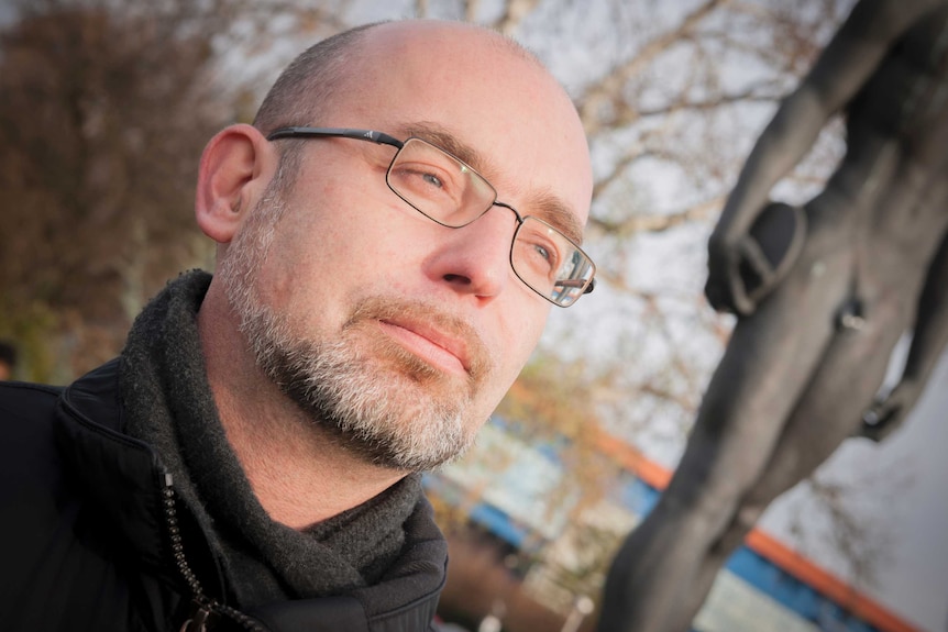 A man with a greying beard and glasses stands near a statue